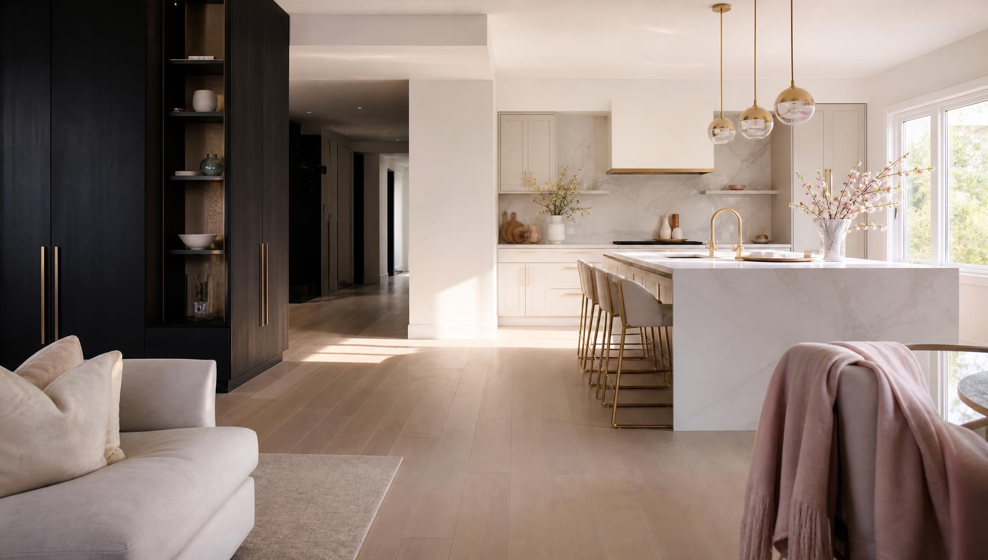 Panoramic spotless open-plan kitchen and living room, dark cabinetry in shadow on the left opening to a sunlit marble island, polished floors and a blush throw