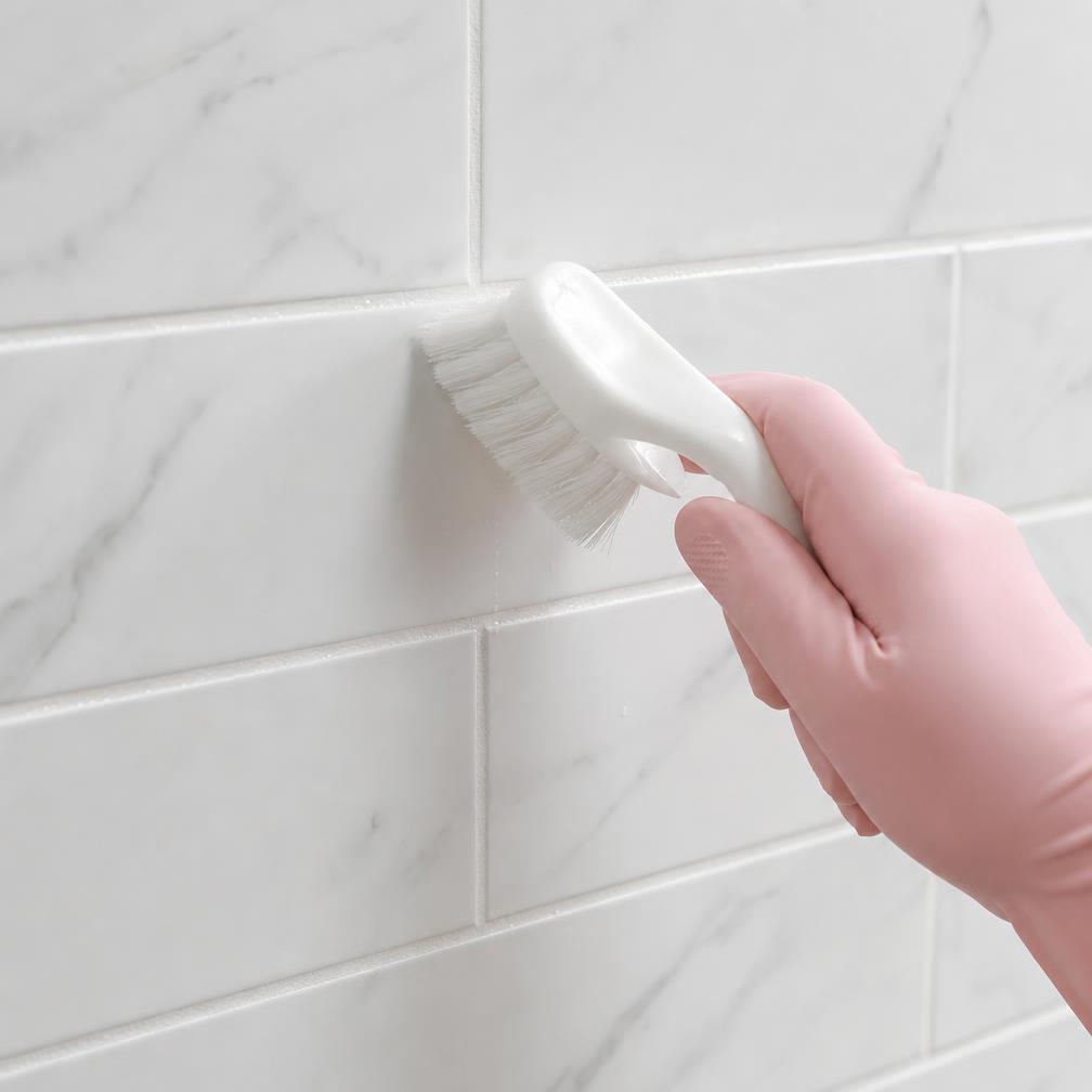 Gloved hand scrubbing white shower tile grout with a grout brush